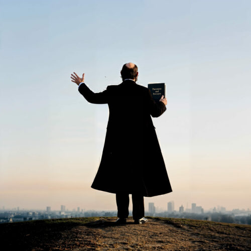 Homme de dos en long manteau noir levant la main droite au sommet d'une colline face à un paysage urbain.