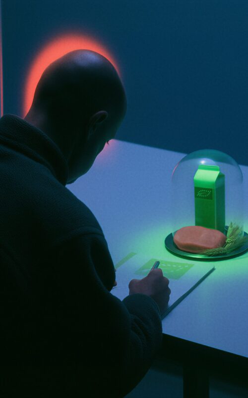 Un homme chauve écrit à une table sombre devant une cloche en verre renfermant une brique de lait et de la nourriture illuminées de vert.