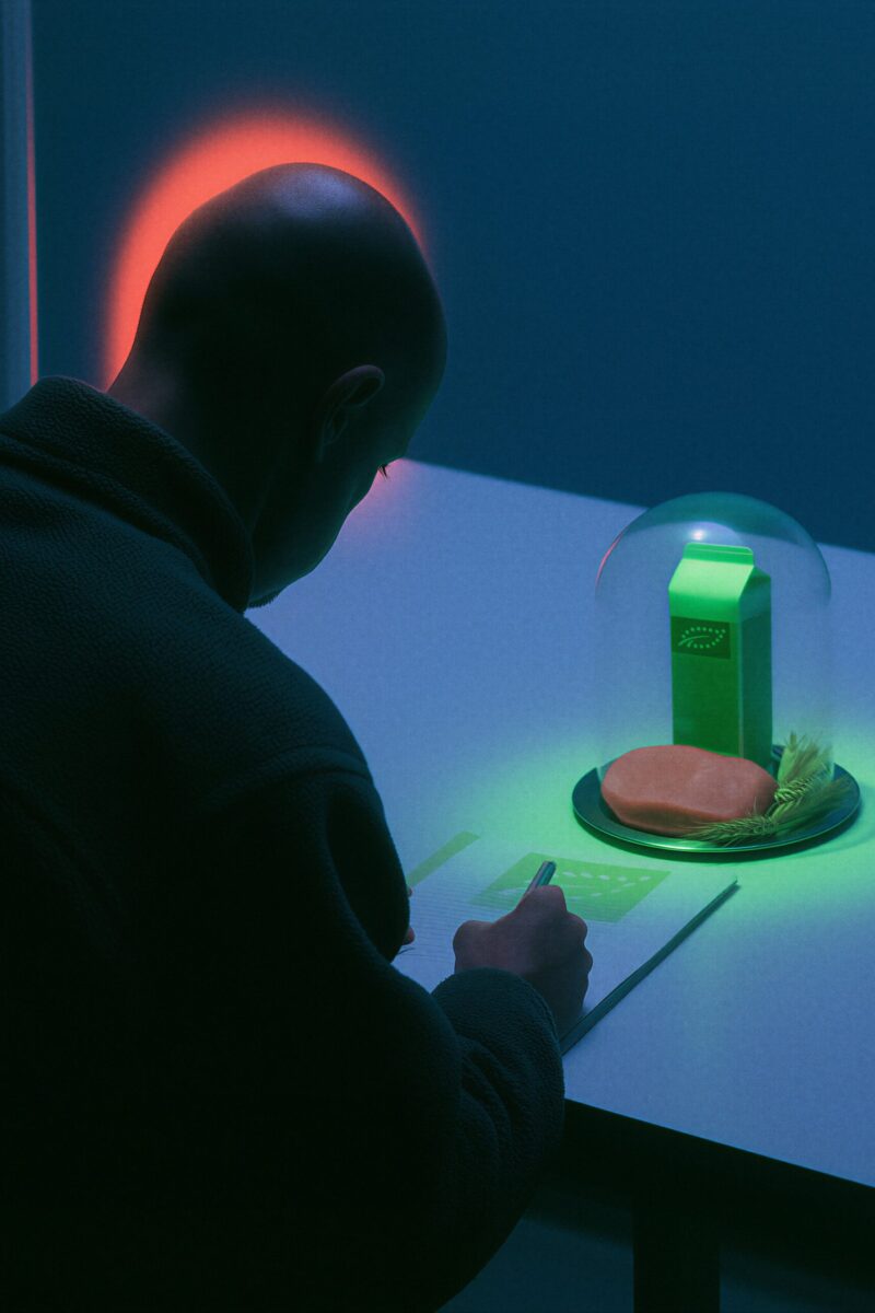 Un homme chauve écrit à une table sombre devant une cloche en verre renfermant une brique de lait et de la nourriture illuminées de vert.