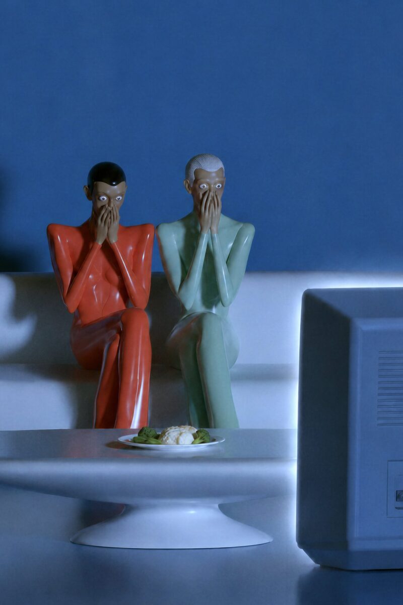 Deux mannequins lisses en tenues moulantes brillantes couvrent leur bouche de stupeur devant la télévision.