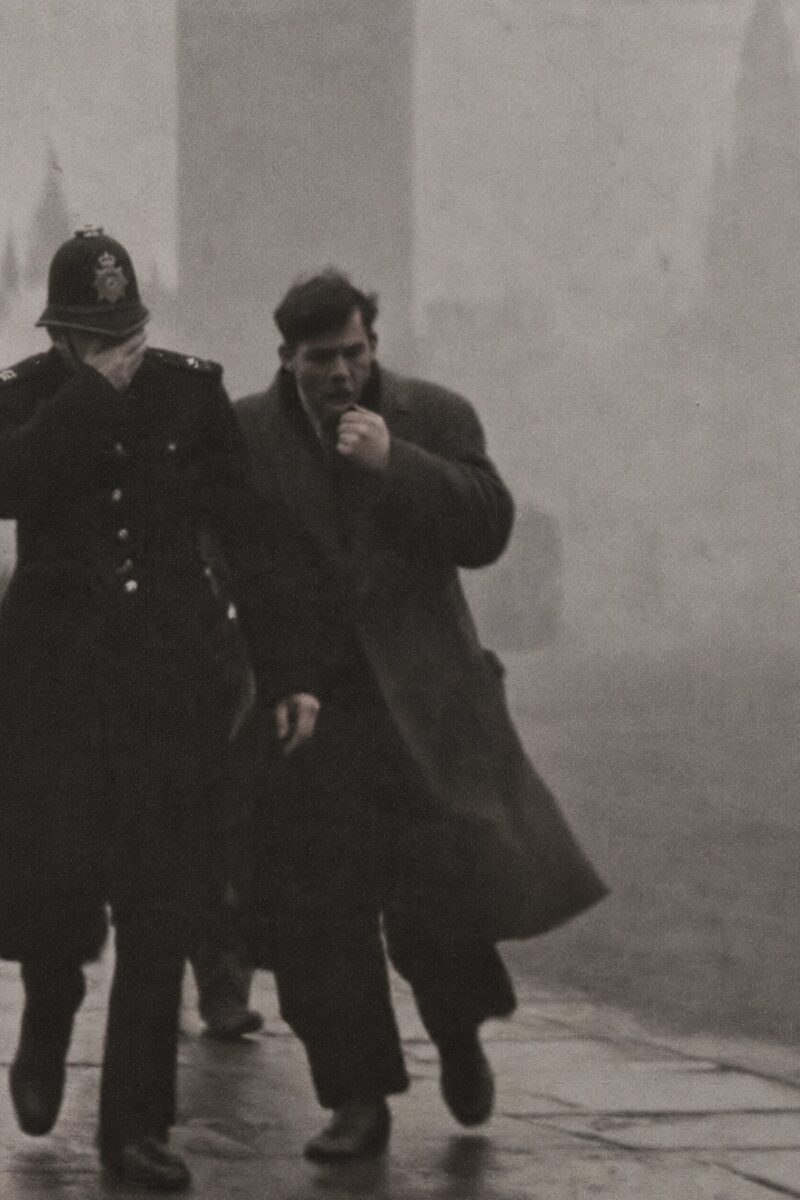 Policier dans le smog londonien Un policier londonien et un passant se couvrent le visage en marchant dans un épais brouillard de pollution.