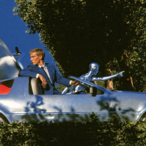 Un homme en costume bleu et un robot humanoïde argenté dans une voiture métallique futuriste.
