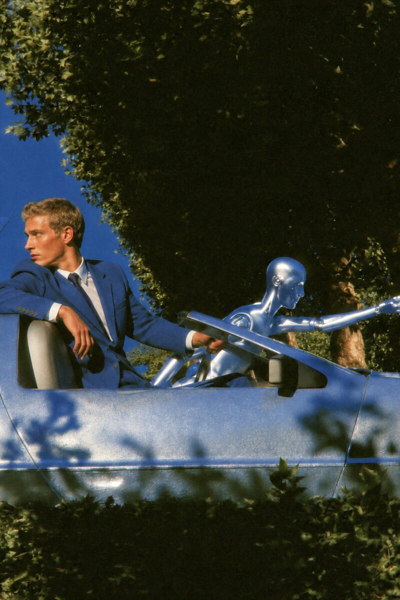 Un homme en costume bleu et un robot humanoïde argenté dans une voiture métallique futuriste.