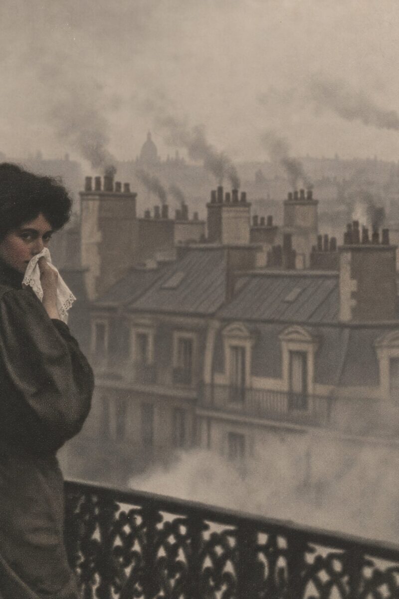Femme vêtue d'une robe d'époque sur un balcon portant un mouchoir à son visage devant les toits enfumés d'une ville.